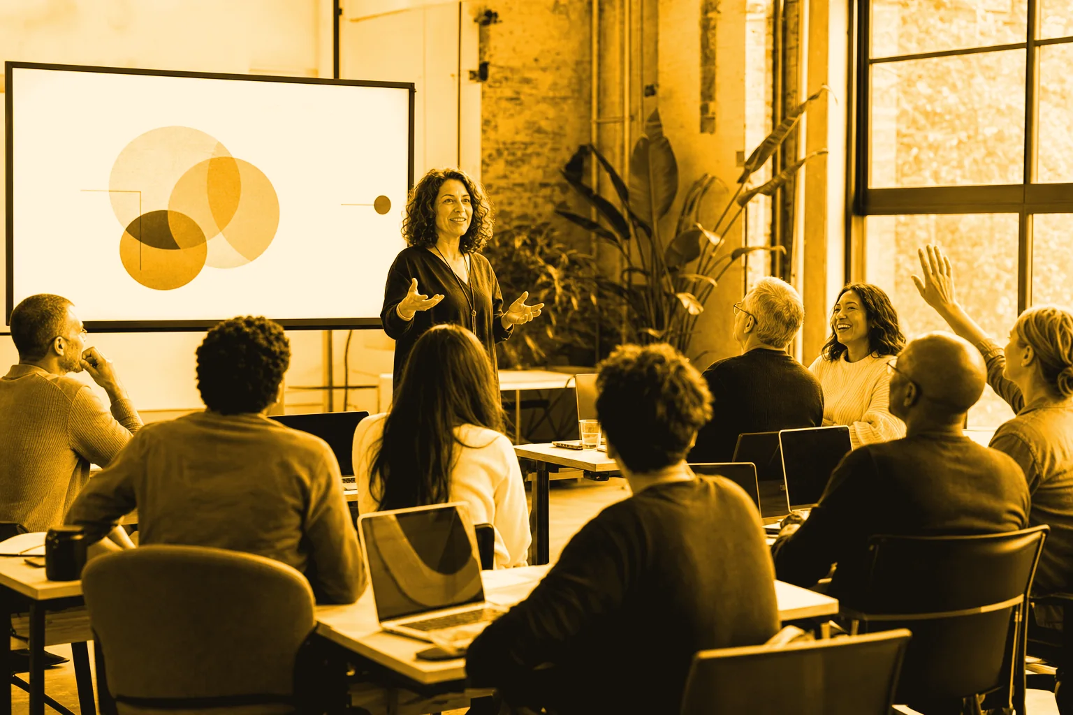 A coach leads a live training session while a room of professionals at laptops works along